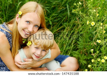 An attractive, smiling mother hugs her child in her lap while sitting outside in the grass and wild flowers