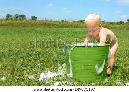 a cute baby boy is outside on a hot summer day, playing in a bucket of water and bubbles