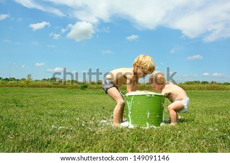 A Young Child And His Baby Brother Are Playing Outside In A Bucket Full Of Bubbles And Water On A Sunny Summer Day