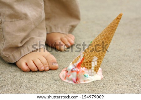 a dropped rainbow colored ice cream cone lays upside down on the sidewalk at the feet of a young child