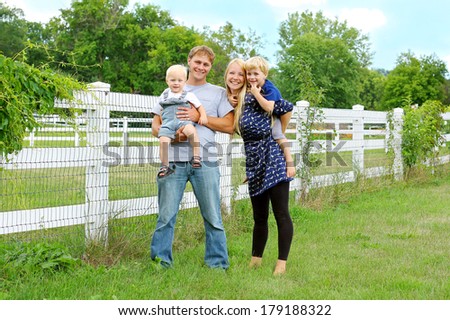 a happy, attractive family of four people, a mother, father, baby and young child are standing outside by a white picket fence on a summer day