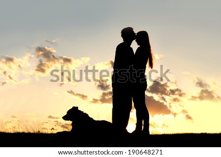 a silhouette of a married couple of man and woman share a loving hug and kiss at sunset, with their German Shepherd Dog laying in the grass