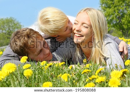A happy family of three people, mother, father, and young child are laying outside in a meadow of dandelion flowers on a spring day.  The little boy is kissing his mom.