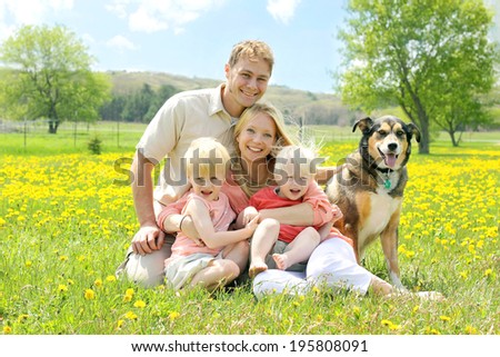Portrait of a happy family of four people, including mother, father, young child, and baby sitting outside with their German Shepherd mix dog on a Spring day.