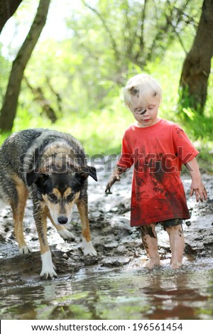 A young boy child and his German Shepherd mix dog are covered in mud and playing outside on the beach of a river in the woods.