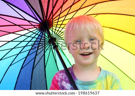 a happy little two year old boy child is smiling as he stands outside under a rainbow colored umbrella to keep dry from the rain.