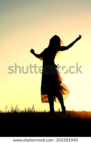 A woman wearing a long skirt, with long blonde hair, is dancing and praising God, while silhouetted against the evening sky