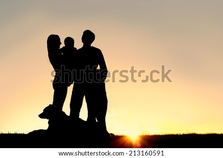 a silhouette of a happy young family of four people; mother, father, child, and baby, stand outside isolated in front of a sunset in the sky with their pet German Shepherd Dog.