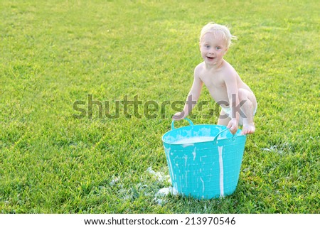 A cute baby boy is making a funny face as he plays outside with bubble soap and water in a wash tub on a summer day.