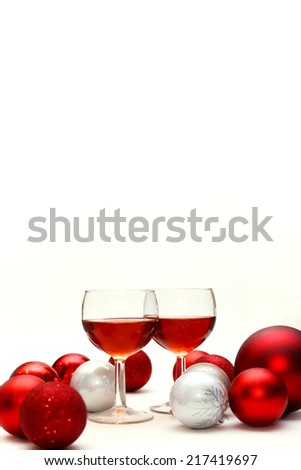 Two wine glasses filled with red wine sit on an isolated white background surrounded by silver and red sparkling Christmas Tree Bulb Decorations.  Copy-space for Greeting Card text or book cover.