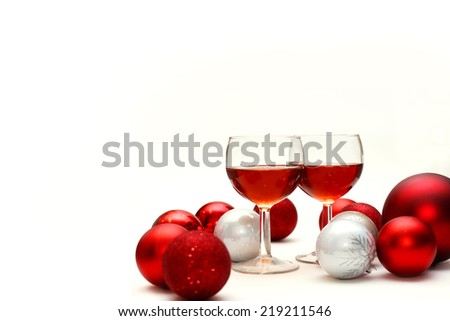 Two wine glasses filled with red wine sit on an isolated white background in surrounded by silver and red sparkling Christmas Bulb Decorations
