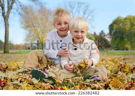 A portrait of two happy, smiling young brothers, a little child and his toddler brother, sitting outside in the fresh fallen Autumn Leaves.