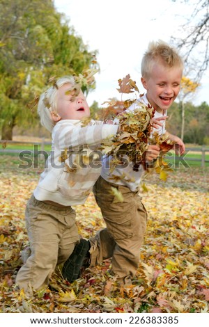 Two young boy children are playing outside on an autumn day, chasing eachother and throwing yellow fall leaves.