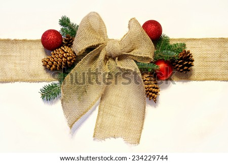 A tan colored burlap ribbon is tied into a bow as if wrapped around a Christmas present, surrounded by natural pine cones, spruce branches and holiday ornaments, and isolated on a white background.