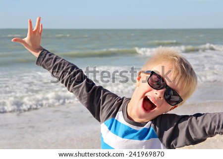 A super happy young child is smiling with joy with his arms raised up while standing on the white sand beach by the ocean on summer vacation.