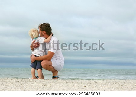 A father and his young toddler son are hugging as they stand on a white sand beach and look out over the ocean while on vacation.