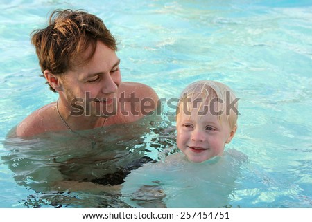 A father is holding his young child in the pool water and teaching him to swim.