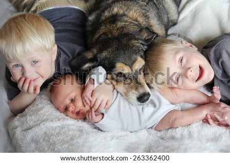 Three happy young children, including a newborn baby girl, a toddler, and their big brother are laying in bed snuggling with their pet German Shepherd Mix dog.