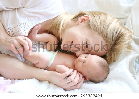 A happy young mother is laying in her white bed, lovingly hugging and snuggling her newborn baby daughter.