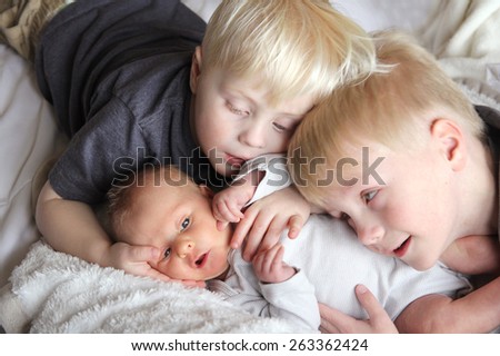 Two young children, a toddler and his big brother are lovingly hugging their newborn baby sister.