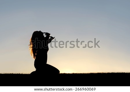 A silhouette of a woman kneeling down with her hands in the air, praying, thanking, and surrendering to God.