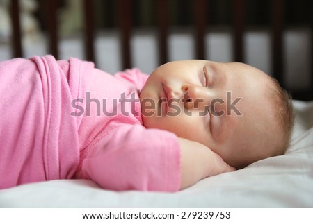 A sweet one month old newborn baby girl is sleeping on her back in her crib, swaddled in a pink blanket.