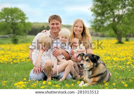 A portrait of a happy family of five caucasian people, including big brother, toddler boy, and baby sister are relaxing in a yellow Dandelion flower meadow with their pet dog.