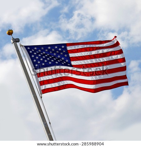 A red, white and blue American Flag is blowing in the wind in front of a cloudy blue sky.