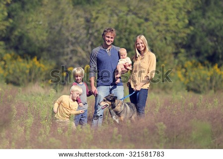 A happy family of 5 people, mother, father with a baby and children, are walking their pet German Shepherd Dog in the meadow on an Autumn day.