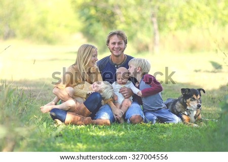 A happy family of five people including mother, father, new baby, boy and his brother are sitting outside in the sunny garden with their adopted German Shepherd dog.  Brother is kissing baby sister.