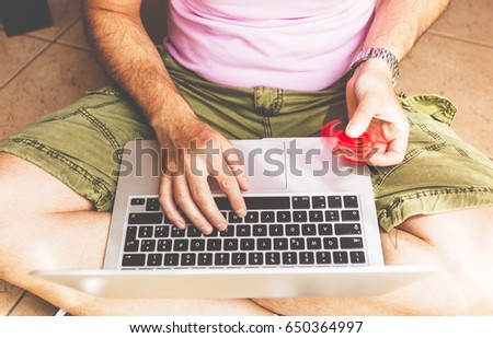 Young adult man seated on a floor, working on laptop and playing with a red hand spinner fidget.