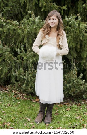 Pretty, young girl warming her hands in a hand muff and standing in front of a fir tree