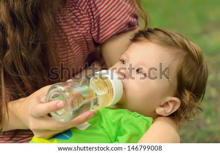 Caring mother gives son drink water bottles on lawn in summer park