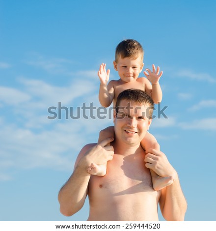 Young father with cute little son sitting on his shoulders on the beach against the sky
