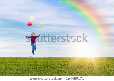 happy young boy levitating with helium balloons against a green meadow and blue sky with a rainbow and a sun burst