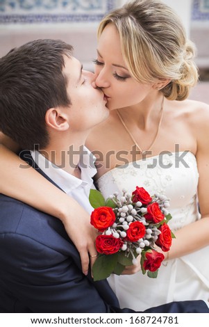Young couple kissing in wedding gown. Bride holding bouquet with red roses