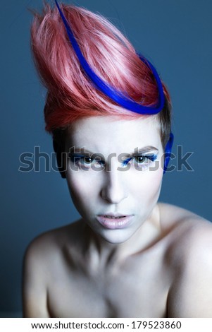 Young person with blue eye shadows, blue ears and pink hair with blue strand on it looking at camera