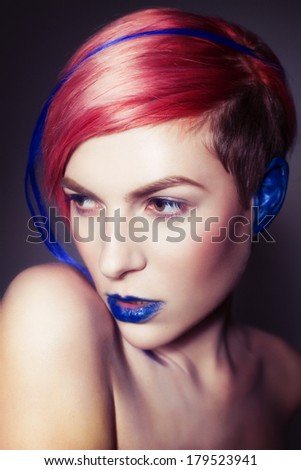 Young person with blue lips, blue ears and pink hair with blue strand on it looking sideways. Blue background