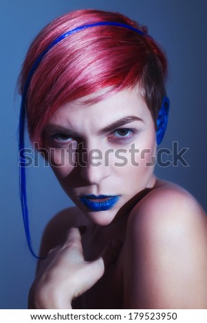 Young person with blue lips, blue ears and pink hair with blue strand on it looking at camera. Blue background