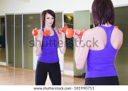 Fit Woman standing with Dumbbells in a Gym looking on herself into a Mirror