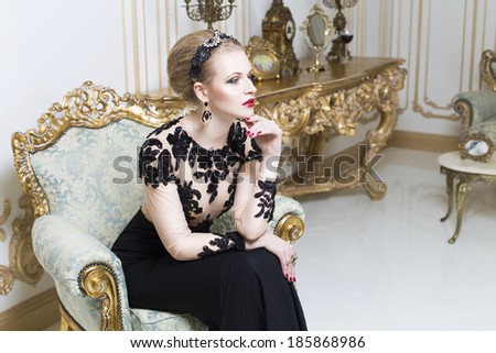 Beautiful blonde royal woman sitting on a retro chair in gorgeous luxury dress. Indoor. Copy Space