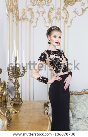 Beautiful blonde royal woman standing near retro table in gorgeous luxury dress. Indoor