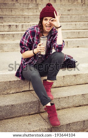 Brunette woman in hipster outfit sitting on steps make fooling gestures on the street. Toned image