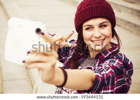 Brunette woman in hipster outfit sitting on steps and taking selfie on retro camera on the street. Toned image
