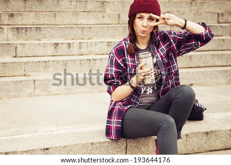 Brunette woman in hipster outfit sitting on steps make fooling gestures on the street. Toned image. Copy Space