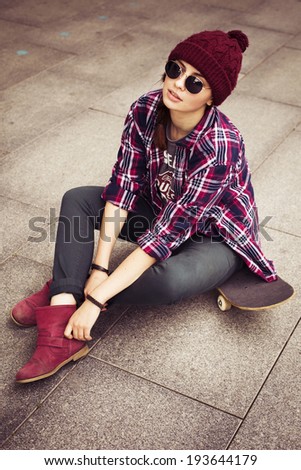 Brunette woman in hipster outfit sitting on a scateboard on the street. Toned image