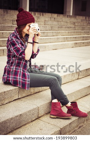 Brunette woman in hipster outfit sitting on steps and photographing on retro camera on the street. Toned image