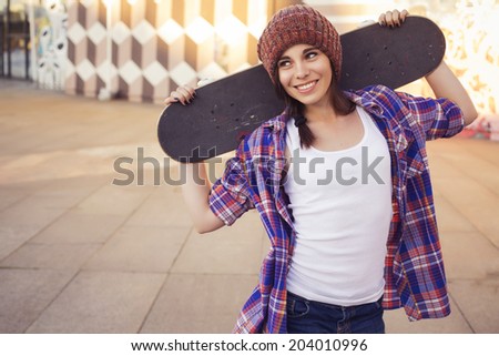 Brunette teenage girl in hipster outfit (jeans shorts, keds, plaid shirt, hat) with a skateboard at the park outdoors. Copy space