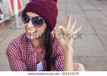 Brunette teenage girl in hipster outfit (jeans shorts, gumshoes, plaid shirt, hat) with a skateboard at the park outdoors.