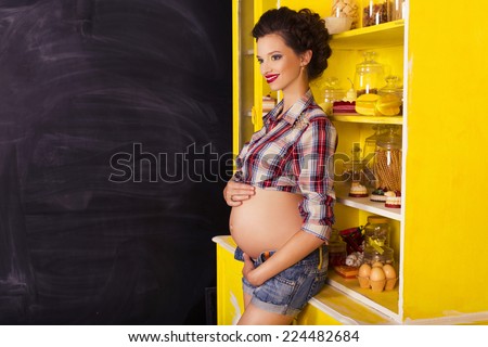 Beautiful brunette woman on a 7th month pregnancy in plaid shirt and jeans shorts on a yellow kitchen and charcoal board. pin up style. copy space